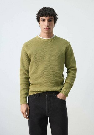 Joven con cabello rizado que lleva un suéter de punto texturizado verde oliva y jeans negros, con una mano en el bolsillo, fondo neutro.