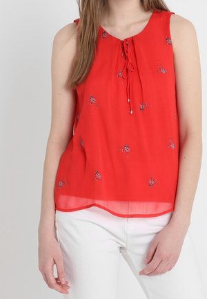 Mouwloze rode blouse met kleine bloemmotieven en strikdetail bij de halslijn, gedragen met witte broek.