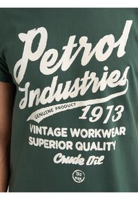 Zelené bavlněné tričko s bílým vintage stylem textu, včetně "Petrol Industries" a "Genuine Product," s prvky jako "1973" a kulatým logem.
