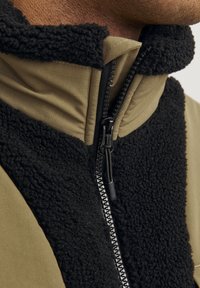 Giacca in pile con colletto alto, materiali a contrasto nero e verde oliva, e una cerniera nera con zip a tirante. Tessuto testurizzato nella parte anteriore.