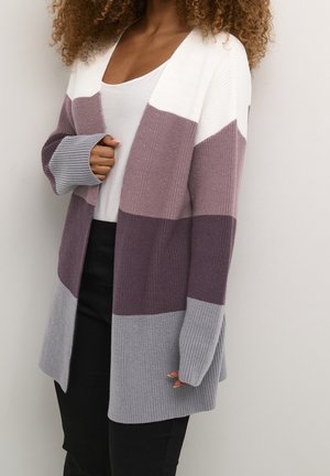Personne portant un cardigan ouvert devant avec de larges blocs de couleurs horizontaux en blanc, mauve, violet et gris sur un haut blanc et un pantalon noir.