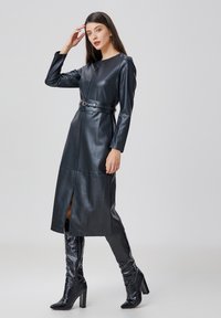 Robe en cuir noir à manches longues, taille cintrée accentuée par une ceinture, et fente à l'avant. Associée à des bottes noires à talons hauts arrivant au genou.