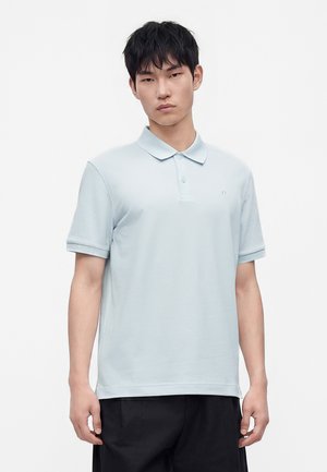 Jeune homme aux cheveux courts noirs portant un polo bleu clair à manches courtes et un pantalon noir, debout devant un fond uni.