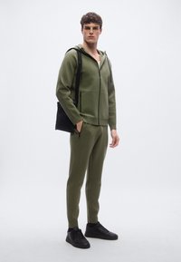 Sudadera con capucha de color verde oliva con cremallera y textura acanalada, combinada con pantalones acanalados a juego, con bolsillos con cremallera y zapatillas negras. El modelo sostiene una bolsa negra.