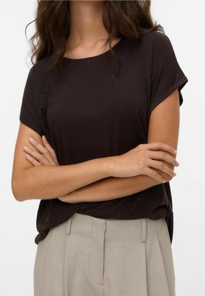 Femme aux longs cheveux bruns, debout les bras croisés, portant une chemise à manches courtes marron foncé et un pantalon beige clair taille haute.