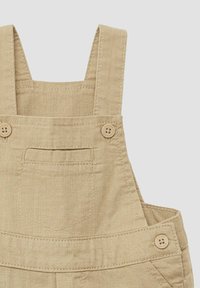 Beige Latzhose mit verstellbaren Trägern, Knopfakzenten und einer aufgenähten Tasche auf der Vorderseite. Der Stoff wirkt strukturiert und strapazierfähig.