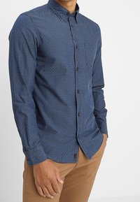 Sjöblå knäppskjorta med små vita prickar, spetsig krage, långa ärmar och en bröstficka. Mjuk tyg med en slät textur.