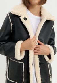 Cappotto in finta pelle nera con dettagli in pelliccia shearling crema, caratterizzato da una chiusura a bottoni, due tasche frontali e un design morbido.