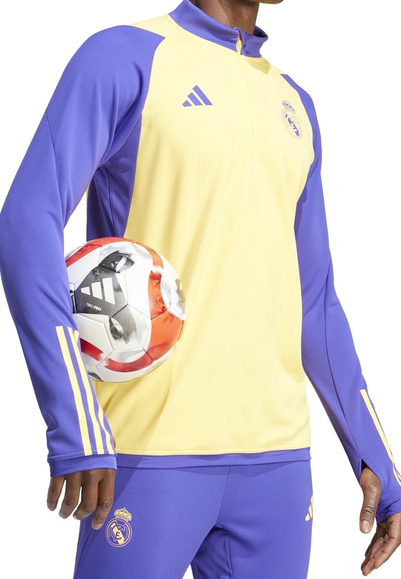 Chándal de Adidas con torso amarillo y mangas moradas, con tres rayas características. Una pelota de fútbol está sostenida bajo el brazo.