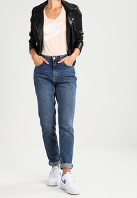 Svart läderjacka för motorcyklister, ljus persiko-färgad Nike-t-shirt med grafik, högmidjade blå jeans och vita sneakers med blå detaljer.