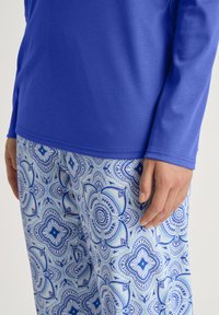 Langärmliges blaues Oberteil, kombiniert mit gemusterten Hosen, die komplizierte Blumenmuster in verschiedenen Blautönen auf einem hellen Hintergrund zeigen.