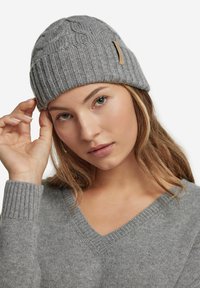 Grå strikket beanie med ribbet kant og kabelmønster, prydet med et lille lædermærke. Blød tekstur, tætsiddende design, ideel til koldt vejr.
