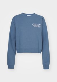 Blå kort sweatshirt med långa ärmar. Har ett vitt tryck med texten "OBEY" och stadnamn. Tillverkad av mjukt material med ribbade muddar.