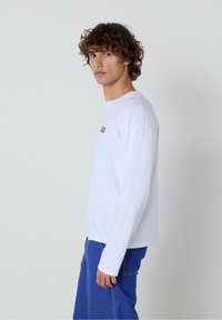 Bijela majica dugih rukava s okruglim izrezom i malim crvenim logotipom na lijevoj strani prsiju, u kombinaciji s plavim trapericama, prikazana bočno.