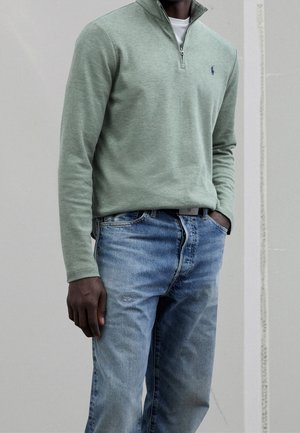 Homme portant un pull vert clair à fermeture éclair quart de zip avec un logo sur la poitrine et un jean bleu délavé, debout avec une main dans la poche contre un mur uni.
