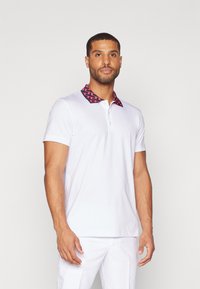 Λευκό πόλο μπλουζάκι με καρό κόκκινο και μαύρο γιακά. Διαθέτει κοντά μανίκια και τριπλή κουμπότρυπα, κατασκευασμένο από ελαφρύ ύφασμα.