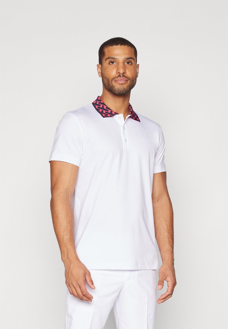 Λευκό πόλο μπλουζάκι με καρό κόκκινο και μαύρο γιακά. Διαθέτει κοντά μανίκια και τριπλή κουμπότρυπα, κατασκευασμένο από ελαφρύ ύφασμα.