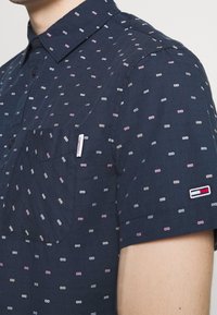 Krátký rukáv námořnicky modré košile s kapsou, zdobené vícebarevným vzorem malých tvarů a značkovým logem na rukávu.