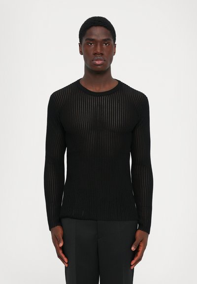 Homme faisant face, portant un pull noir à manches longues ajusté et des pantalons noirs, sur un fond clair.