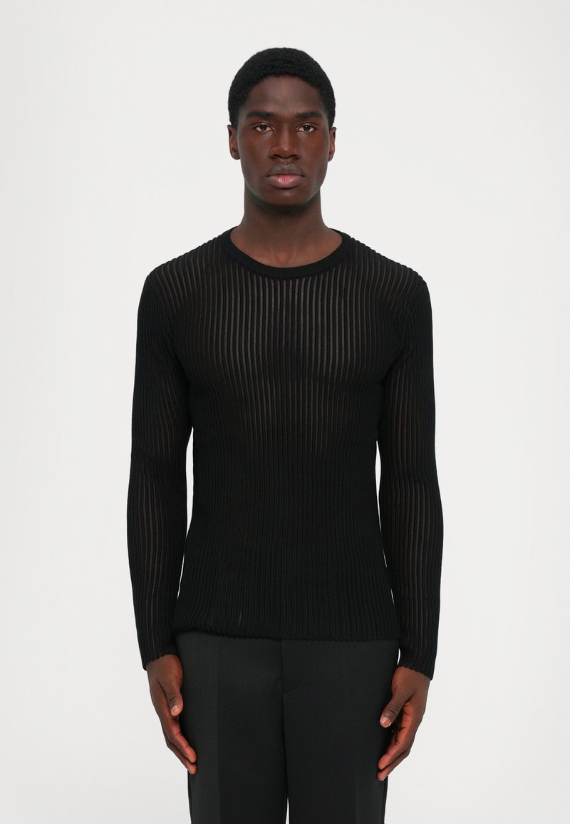 Άνδρας που κοιτάζει μπροστά, φορώντας ένα εφαρμοστό μαύρο μακρυμάνικο ριμπέ πουλόβερ και μαύρο παντελόνι, μπροστά σε απλή ανοιχτόχρωμη φόντο.