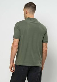 Mann iført olivengrønn kortermet poloshirt og svarte jeans, sett bakfra mot en ensfarget lys bakgrunn.