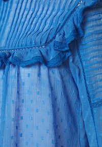 Sinine kleit, millel on tekstuuriga, poolläbipaistev kangas, rippuv pitsääris ja väikesed, kõrgendatud täppmustrid kogu ulatuses.