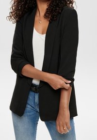 Blazer nero con una vestibilità su misura, rever strutturati, maniche arrotolate e un tessuto liscio, abbinato a una maglietta bianca e jeans blu.
