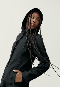 Črna kapuča s prilegajočim dizajnom, stranskimi žepi in gladko teksturo. Ima dolge pletene lase in enostavno ozadje.