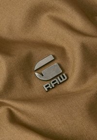 Metallogo som läser "RAW" på texturerad beige tyg, med en slät design och en böjd form med skarpa kanter.