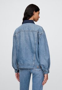 Svetlo modra denim jakna z ohlapnim pristopom, ki ima temen ovratnik, dolge rokave in dva zadnja žepa z kovinskimi poudarki.