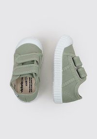 Baskets en toile vert clair avec semelles en caoutchouc blanches et deux bandes Velcro. Elles comportent une languette avec une marque à l'intérieur.