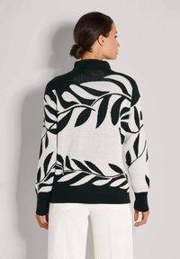 Frau, die einen schwarz-weißen Pullover mit Blattmuster und weiße Hosen trägt, von hinten gegen eine schlichte Wand betrachtet.