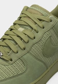 Baskets vert olive avec des panneaux en daim et en maille, des lacets épais assortis et un logo subtil en relief en forme de swoosh sur le côté.