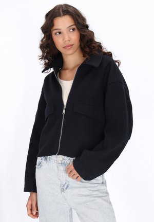 Giacca nera corta con zip frontale, ampio colletto e due tasche frontali, abbinata a jeans azzurri a vita alta. Sfondo neutro.