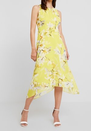 Ärmelloses gelbes Kleid mit weißem und grauem Blumenmuster, asymmetrischem Saum, getragen mit weißen Absatzschuhen mit Knöchelriemen.