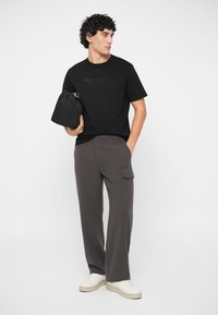 Uomo che indossa una t-shirt con logo nero, pantaloni cargo grigi, scarpe da ginnastica bianche, con una mano nella tasca e l'altra che tiene una borsa nera, mentre guarda di lato.