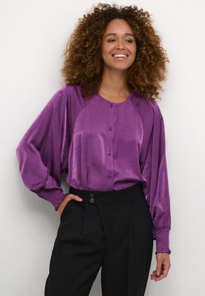 Blusa di satin viola con frontale a bottoni, maniche raglan e polsini elasticizzati, abbinata a pantaloni neri a vita alta.