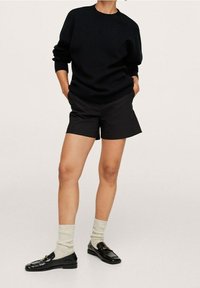 Schwarzer Strickpullover, schwarze Shorts, graue gerippte Socken und schwarze Slipper mit goldenen Akzenten. Lässiges Outfit mit einem einfachen, klaren Design.