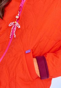 Dłoń w żółtej rękawicy spoczywa w kieszeni pikowanej, jasnopomarańczowej kurtki z fioletowym i różowym zamkiem błyskawicznym oraz koralikowym uchwytem zamka.