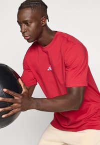 Sarkana īsām piedurknēm kokvilnas T-krekls ar apaļu kakla izgriezumu un baltu Adidas logotipu. Personas rokās ir melna medicīnas bumba, kas izstaro koncentrāciju darbībā.