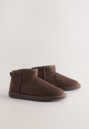 Braune Wildleder-Stiefeletten mit weicher Textur, abgerundeten Zehen und minimalen Nähten. Flache Gummisohlen mit einer Zuglasche hinten.