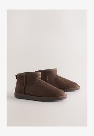 Braune Wildleder-Stiefeletten mit weicher Textur, abgerundeten Zehen und minimalen Nähten. Flache Gummisohlen mit einer Zuglasche hinten.