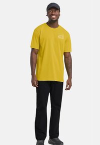 Gelbes T-Shirt mit kurzen Ärmeln und kleinem Aufdruck auf der Brust, kombiniert mit schwarzer Hose. Modell trägt eine schwarze Mütze und steht vor einem schlichten Hintergrund.