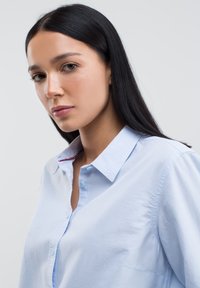 Camicia button-up di cotone azzurro chiaro, con colletto, texture sottile e un accento rosso sotto il colletto.