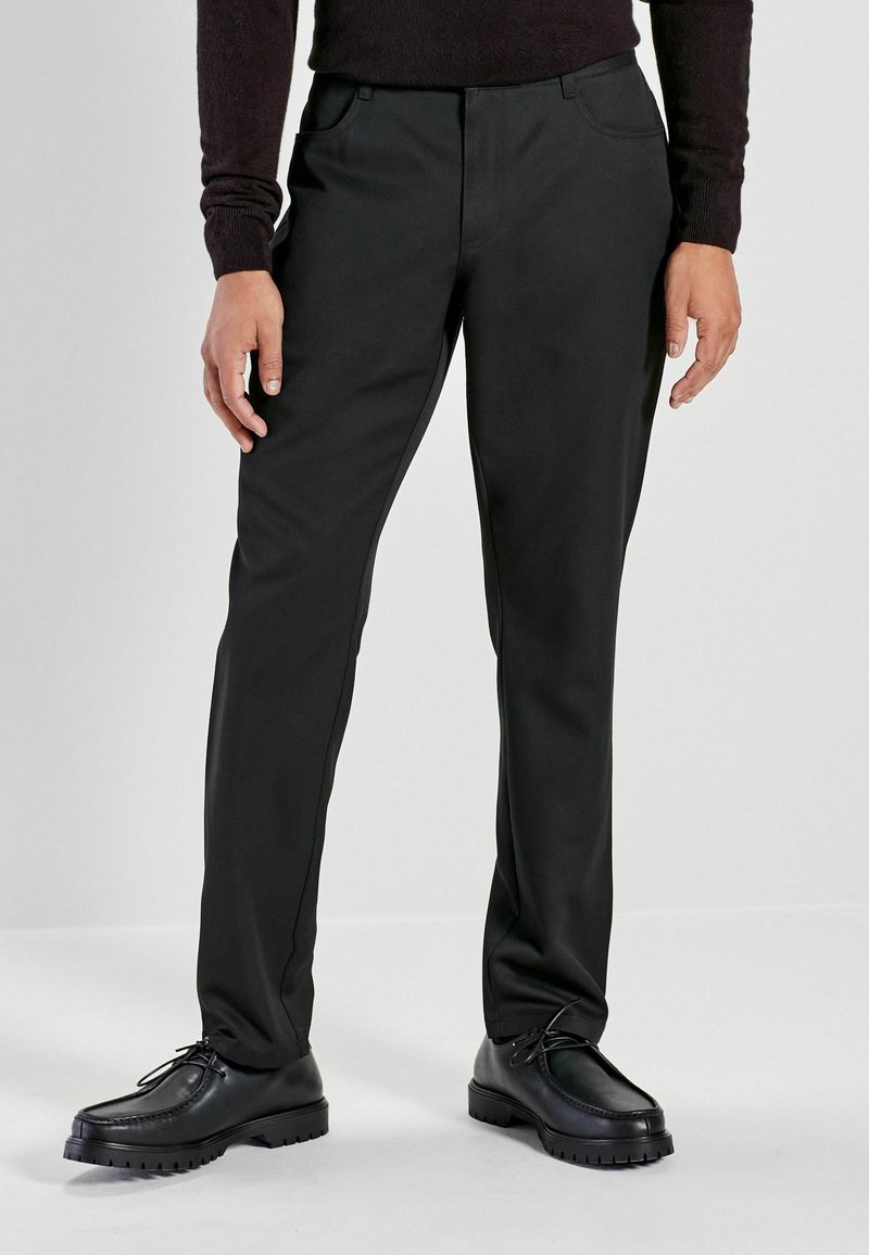 Black trousers with a smooth texture, standard fit, two front pockets, and contrasting seams. Paired with black shoes featuring a rugged sole.