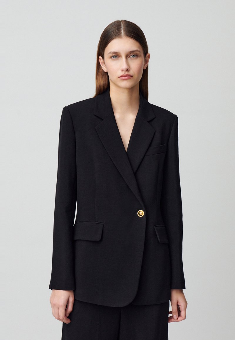 Blazer negro de tela lisa, con un solo botón dorado, solapas con muesca, dos bolsillos frontales, corte ajustado, mangas largas, diseño minimalista.