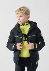 Chaqueta acolchada negra con detalles amarillos, bolsillos con cremallera y capucha. Llevada sobre un suéter amarillo brillante con un diseño de logo negro.