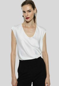 Blusa bianca a maniche corte con scollatura a V, realizzata in tessuto morbido, abbinata a pantaloni neri su misura, che mostrano un look minimalista.