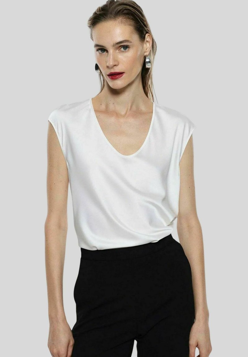 Blusa bianca a maniche corte con scollatura a V, realizzata in tessuto morbido, abbinata a pantaloni neri su misura, che mostrano un look minimalista.