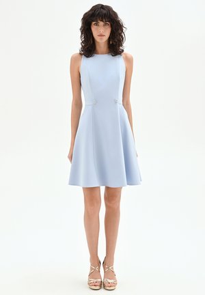 Femme aux cheveux bouclés et foncés portant une robe trapèze sans manches bleu clair et des sandales à talons hauts dorées à lanières, sur fond blanc.